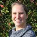 Portrait von Daniela Scheiler in einem grauen Strickpullover, die warm in die Kamera lächelt mit grünem Laub im Hintergrund.