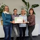Vier lächelnde Frauen stehen zusammen und halten ein gerahmtes Zertifikat. Grüne Pflanzen sind im Hintergrund vor weißer Wand sichtbar.