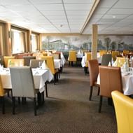 Interior of MS De Willemstad ship restaurant with colorful upholstered chairs, white tablecloths, and historic Dutch canal scene murals on walls.