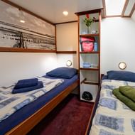 Compact cabin on ship Elizabeth with two single beds with blue pillows and patterned bedding, burgundy carpet, and shelving unit between beds.