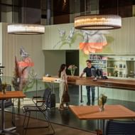 Moderne Hotellobby mit Holzempfangstresen, floraler Wandkunst, runden Kronleuchtern und Gästen am Tresen. Holztische mit gestreiften Stühlen im Vordergrund.