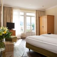 Helles Hotelzimmer im Hotel Seeburg Luzern mit zwei Einzelbetten, Holzschrank, TV und Balkontür mit Blick auf See und Berge.