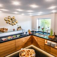 Breakfast buffet with bread rolls, cold cuts, cheese, and fresh items on wooden counters with glass shields at Gartenhotel Heusser.