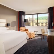 Modern superior room at Sheraton Essen with white double bed, orange patterned armchair, desk, and large window offering city views.