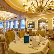 Elegant hotel restaurant with white-clothed tables, wicker chairs, ornate chandeliers, and arched mirrors. Warm golden lighting and decorative ceiling.