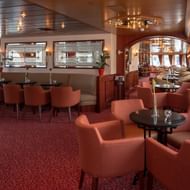 Elegantes Bar-Interieur auf der MS Switzerland mit korallfarbenen Sesseln, runden Tischen, rotem Teppich und holzgetäfelter Decke mit Beleuchtung.