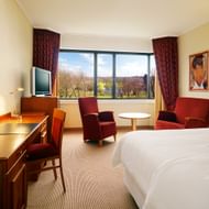 Bright hotel room at Sheraton Essen with double bed, desk, red armchairs, TV, and large window overlooking a park with autumn trees.