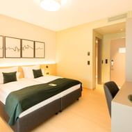 Modern hotel double room with white bedding, green runner, desk with lamp, and abstract wall art. Warm lighting creates a welcoming atmosphere.