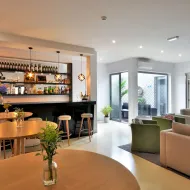 Modern hotel bar with wooden tables, bar stools, backlit shelves with bottles, and lounge seating with beige and green armchairs at Porto Domus Hotel.