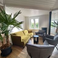 Bright lounge at Backbord Hotel with yellow sofa, gray armchairs, palm plants, and white paneled ceiling with window view.