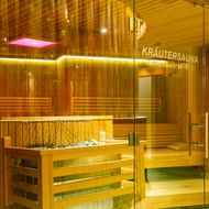 Modern herbal sauna with wooden benches, glass door, warm lighting and pink infrared panel. Sign reads 'Kräutersauna' with temperature 60-65°C.