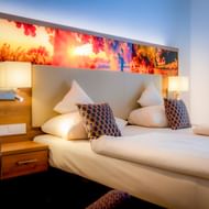Modern double room with white bedding, decorative pillows, wooden nightstands, reading lights, and colorful backlit headboard artwork.