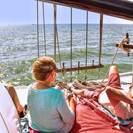 Two people relaxing on red cushions on the deck of sailing ship Elizabeth, with ropes and rigging visible, overlooking calm sea waters.