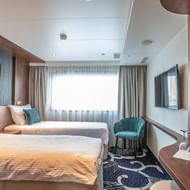 Modern MS Swiss Crown twin cabin with two beds, teal chair, wall-mounted TV, large windows with curtains, and contemporary decor in neutral tones.