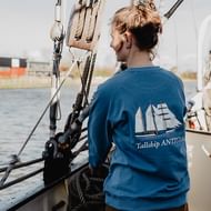 Crewmitglied in blauem Tallship Antigua Sweatshirt steht an Deck und arbeitet mit Takelage und Seilen. Hafengebäude im Hintergrund sichtbar.