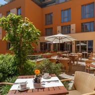 Outdoor breakfast terrace at Best Western Bamberg with set tables, wicker chairs, white umbrellas, and garden. Orange hotel building in background.