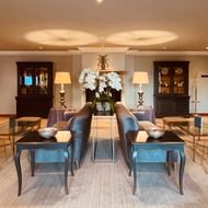 Elegante Hotellobby mit blauen Samtsesseln, beigen Sesseln, Glastischen und weißer Orchidee als Mittelpunkt. Warme Beleuchtung und dunkle Holzschränke.