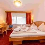 Bright hotel room with wooden furniture, double bed with white pillows, red curtains, desk with TV, and red carpet at Hotel Schütthof.
