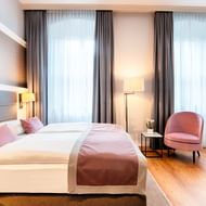Modernes Hotelzimmer im Welcome Residenzschloss Bamberg mit zwei Einzelbetten, rosa Sessel, Schreibtisch und großen Fenstern mit grauen Vorhängen.
