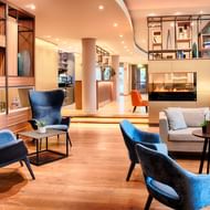 Modern Welcome Hotel Marburg lobby with blue velvet armchairs, gray sofa, wooden shelving, fireplace, and warm lighting throughout the space.