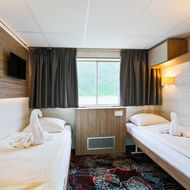 Compact cabin on MS Normandie with two single beds, white linens, wood paneling, TV, window with curtains, and patterned carpet.
