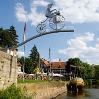 Werra-Radweg Skulptur in deutscher Stadt Metallskulptur eines Radfahrers auf Hochrad über der Werra. Historische Fachwerkhäuser und Steinmauer am Flussufer sichtbar.