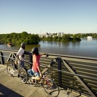 Zwei Radfahrer mit Fahrrädern stehen am Brückengeländer mit Blick auf die Elbe in Hamburg, mit Hafenszenerie und grünen Ufern.