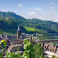 Panoramic view of Bernkastel-Kues with historic buildings, church spire, bridge over Moselle River, vineyard slopes and forested hills.