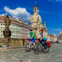 Zwei Radfahrer mit Tourenrädern und Packtaschen auf Kopfsteinpflaster in Dresden vor der barocken Frauenkirche-Kuppel und historischen Gebäuden.