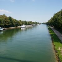 Ems-Kanal mit Radweg und Schiffen Breiter Kanal mit grünem Wasser, gesäumt von Bäumen. Boote am linken Ufer vertäut, gepflasterter Radweg rechts mit Radfahrern in der Ferne.