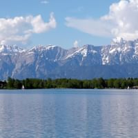 Zeller See mit schneebedeckten Alpengipfeln im Hintergrund, grünem Waldufer und kleinen Segelbooten auf ruhigem blauen Wasser unter bewölktem Himmel.