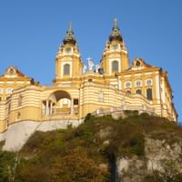 Gelbes Barockkloster mit Zwillingstürmen und grünen Kuppeln auf einem felsigen Hügel vor klarem blauen Himmel, umgeben von Vegetation.