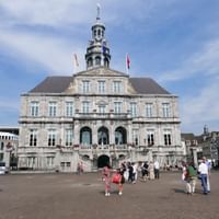 Rathaus Maastricht, ein barockes Steingebäude mit verziertem Turm und Uhr, wehende Flaggen. Touristen versammeln sich auf dem Platz davor.