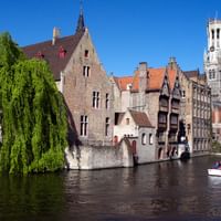 Medieval buildings line a canal in Bruges with the famous Belfry tower visible. A weeping willow hangs over the water and a small boat carries tourists.