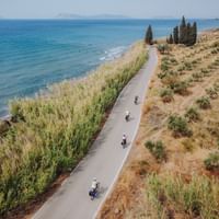 Küsten-Radweg auf dem Peloponnes, Griechenland Luftaufnahme von Radfahrern auf einem asphaltierten Küstenweg auf dem Peloponnes, Griechenland, zwischen türkisfarbenem Meer und mediterraner Vegetation.