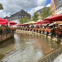 Stone-walled canal flowing through Saarburg old town with red umbrellas, flower boxes, and historic buildings lining both sides.