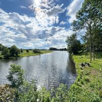 Dove Elbe river flowing through green landscape near Geesthacht. People rest on grassy riverbank under trees, with blue sky and white clouds above.