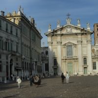 Mantua Cathedral with ornate baroque facade and bell tower on cobblestone square. Historic buildings line the plaza with visitors walking around.