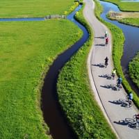 Luftaufnahme von Radfahrern auf einem gewundenen Radweg durch grüne Nordholland-Landschaft mit Wasserkanälen zu beiden Seiten.
