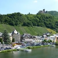 Bernkastel-Kues an der Mosel mit historischen Gebäuden, Parkplatz, Schiffsanleger, weinbergbedeckten Hügeln und Burgruine auf dem Hügel.