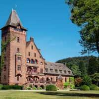 Rotes Backsteinschloss in Mettlach mit konischem Turm, umgeben von grünen Rasenflächen und alten Bäumen unter blauem Himmel mit bewaldeten Hügeln.