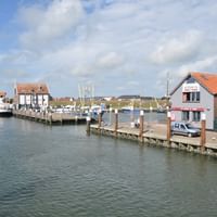 Harbor view of Oudeshild with wooden pier, moored boats, and traditional Dutch buildings with red tile roofs under a cloudy blue sky.