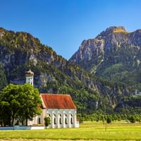 White church with red roof and bell tower in green meadow near Schwangau, with Neuschwanstein Castle visible on forested mountain slopes.