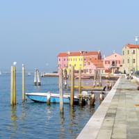 Blick auf die Uferpromenade von Pellestrina mit bunten Häusern in Rosa, Gelb und Orange entlang des Kanals. Hölzerne Anlegepfähle und kleine Boote.