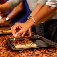 Person decorating chocolate on a tray with cocoa beans scattered on the work surface during a chocolate making workshop.