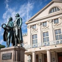 Bronzedenkmal von Goethe und Schiller vor dem klassizistischen Deutschen Nationaltheater in Weimar unter blauem Himmel.