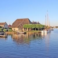 Charming Dutch cottages with thatched roofs line a peaceful waterway in Holland, with sailboats moored at wooden docks under a clear blue sky.