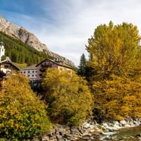 Herbst im romantischen Splügen Alpendorf Splügen mit traditionellen Holzchalets, weißem Kirchturm und goldenen Herbstbäumen entlang eines Bergbachs.