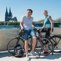 Man and woman with touring bikes sitting on Rhine Boulevard in Cologne with cathedral and Hohenzollern Bridge in background.