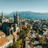 Aerial view of Lausanne old town with cathedral towers, historic buildings, and Lake Geneva with mountains in the background under clear sky.